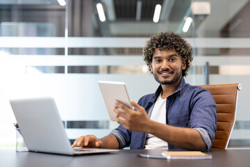 Portrait of a young Indian man working in an office at a laptop, holding a tablet in his hand and looking at the camera with a smile