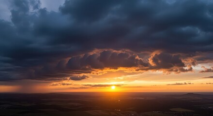Intense golden sunset illuminates dramatic dark storm clouds hovering over a vast landscape below