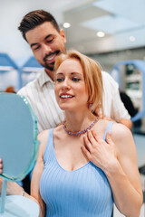 Happy couple shopping for jewelry in a luxury store, with the man lovingly placing a stunning necklace around his smiling blonde girlfriend's neck, celebrating their romance