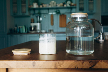Jar of sourdough starter next to water jug on wooden kitchen table  