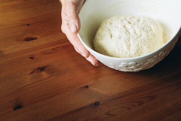 Hand holding bowl of sourdough dough on wooden table in kitchen  
