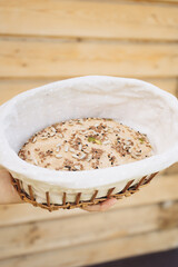 Sourdough with seeds in proofing basket held by person on wooden background  