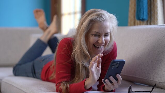 A woman with long blonde hair waving and smiling using smartphone for video chat with friends is lying on a white couch, wearing a red sweater and jeans.