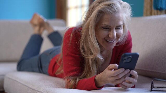 Smiling mature woman is comfortably lying on a couch as she texts on her smartphone. The space has a bright, vibrant atmosphere with colorful decor and sunlight streaming in. - Powered by Adobe
