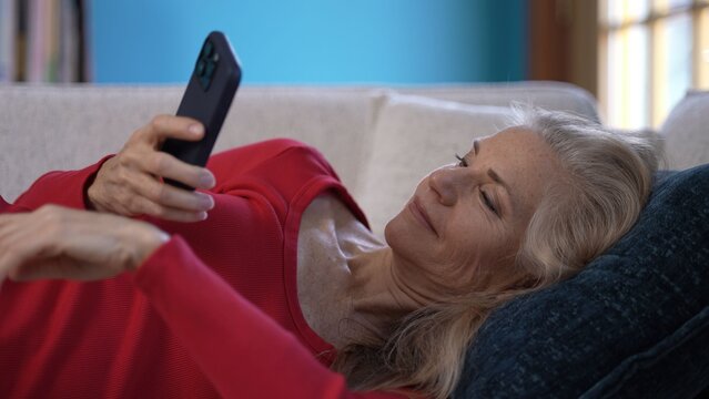 Mature senior woman lounges comfortably on a couch, using her smartphone. She enjoys a peaceful moment in well lit living room filled with books and calming atmosphere. - Powered by Adobe