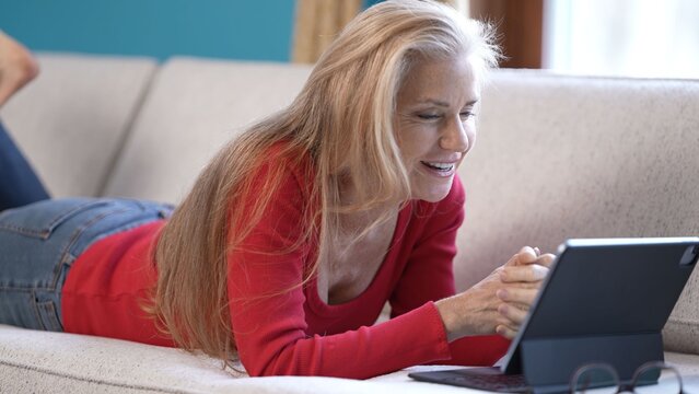 A mature woman lounges comfortably on a couch, smiling as she reads good news on her tablet. Relaxed atmosphere shows her enjoyment during a leisurely moment in living room.