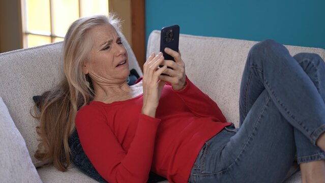 Angry frustrated mature woman relaxes on a couch, wearing a red top and jeans. She looks surprised while looking at her smartphone, and gets her attention in a bright living room