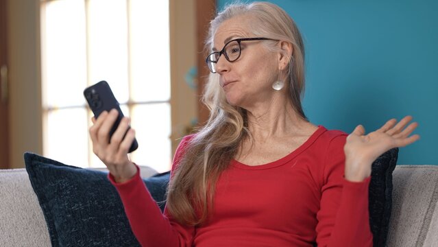 Unhappy stressed middle-aged woman in a red sweater sits on a couch, carefully reading a letter. She appears deep in thought, surrounded by a cozy home environment.