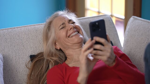 A middle-aged woman lies comfortably on a couch, smiling widely as she interacts with her smartphone, enjoying a moment of joy and relaxation in a cozy room.