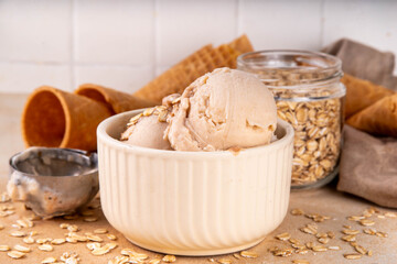 Vegan oats milk non-dairy ice cream, scoops of vanilla creamy oatmeal milk gelato in beige bowl, with ice cream waffle cones and rolled oats flakes