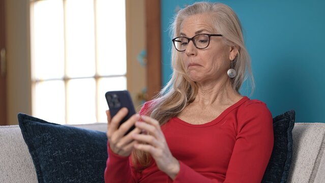 Mature woman with long, silver hair sits comfortably on a cozy couch, beams with joy while holding her smartphone. She expresses delight as she engages in a heartfelt conversation.