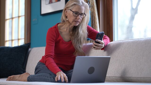 A woman with blonde hair and glasses, waving at phone having video chat, sits on a couch with a laptop and a phone. She is smiling and waving her hand.