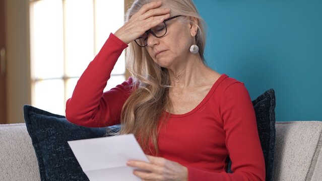 Frustrated middle-aged woman with long hair reads a bad news letter with an expression of surprise and concern. She sits comfortably on a sofa against a bright blue wall.
