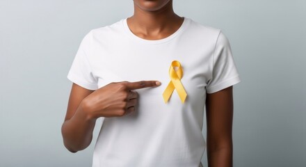 African American woman stands with a yellow ribbon on a white t-shirt, pointing at it. Banner template for childhood cancer awareness month concept and support for disease patients.