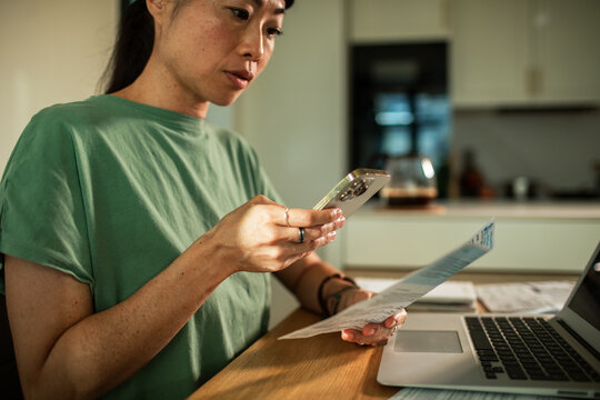 Adult woman focused scanning bill with smartphone at home kitchen table