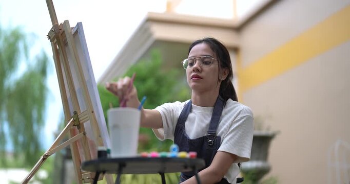 asian woman sitting in front of easel painting with gentle smile during creative afternoon in backyard using brush to create calm landscape artwork enjoying quiet hobby time in natural garden space