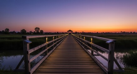 Obraz premium Long wooden footbridge leads toward a distant structure under a vibrant twilight sky