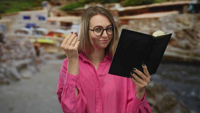 Woman in pink shirt reading book at beach, gesturing money sign, with blurred seaside background, conveying financial independence and relaxation outdoors.