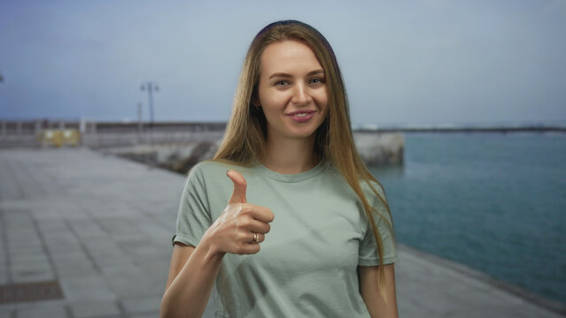 Woman smiling on promenade near sea gives thumbs up on a bright day, showcasing joy and positivity with a serene ocean backdrop emphasizing leisure and relaxation.