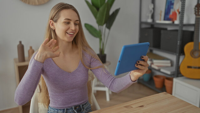 Young woman in casual clothes using tablet at home in bright living room engaging in video call with modern decor and plants visible in the background.