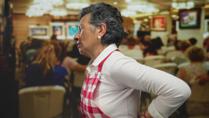 Obraz premium Senior woman with grey hair in apron at art gallery indoor setting, surrounded by seated people, appears focusing intently on invisible artwork display.
