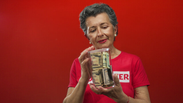 Senior woman with grey hair holds money jar while volunteering against a vibrant red background, showcasing charity and financial support themes.