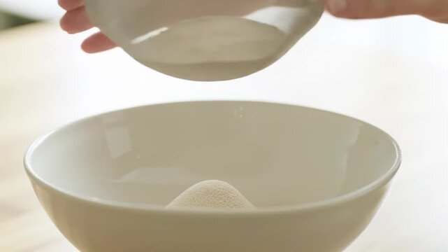 Clean hands gently sift light beige powder into a white ceramic bowl on a wooden countertop, showcasing exquisite texture in ultra-slow-motion with soft natural daylight and creamy bokeh. Concept of
