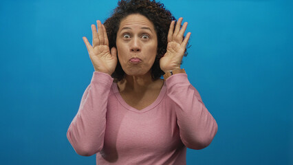 Middle aged hispanic woman covering face with hands in blue studio then uncovering while smiling and waving; joy playful gesture.