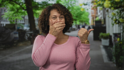 Middle-aged hispanic woman wearing pink top covers mouth with hand and holds thumb up on busy city...