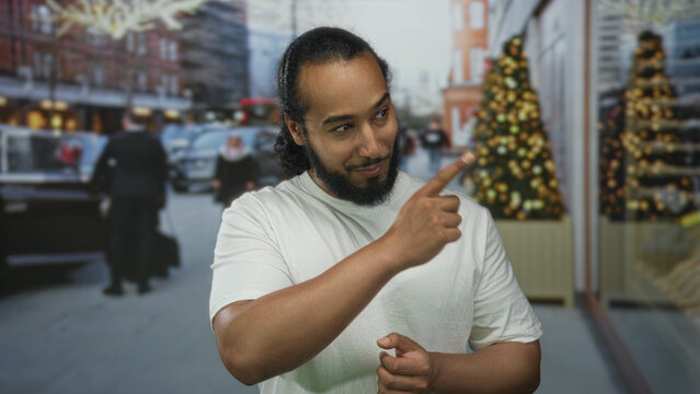 Bearded man in white tshirt points finger to shop window on busy city street with christmas trees and passing shoppers; friendly greeting.