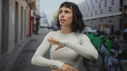 Hispanic woman gestures with raised hands while talking on a busy urban city street during daylight; assertion communication confidence.