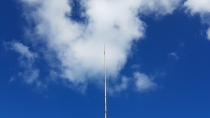 Striking view of a tall communication tower reaching high into the clear blue sky, piercing through a fluffy white cloud formation