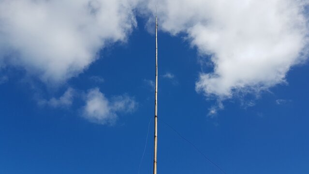 Low angle perspective of a slender transmission tower reaching up into the vast blue sky between two white clouds