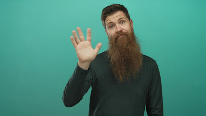 Man with long brown beard raises bare hand with open palm against a teal studio background; curiosity.