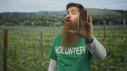 Fototapeta premium Man wearing green volunteer shirt raises hand in stop gesture in a countryside field; safety awareness.