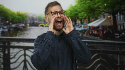 Man with glasses and beard cupping hands to mouth on a street canal bridge in amsterdam near cafe...