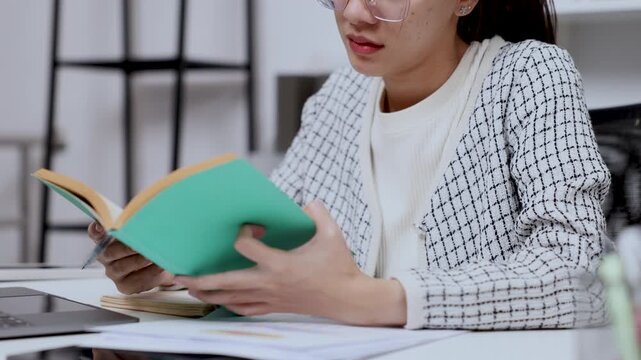 asian female entrepreneur reading book with interest while sitting at home office desk researching idea for business strategy development planning and skill enhancement