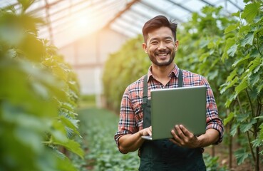 Asian farmer in greenhouse holds laptop checking plant growth. He smiles analyzing farm data with technology for better yield. Cultivation success in modern agriculture.