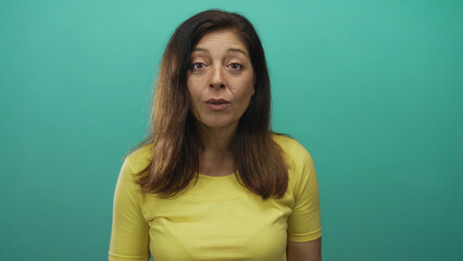 Fototapeta premium Middle aged hispanic woman with hands on head in studio environment with teal background and yellow shirt; surprise.