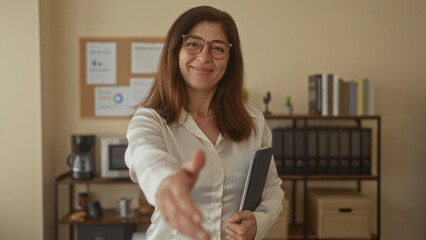 Woman extends hand for handshake holding tablet in office by bookshelf and bulletin board with a bright smile  friendly welcome. © Krakenimages.com