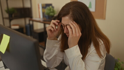 Middle aged woman wearing glasses sitting at computer resting hand on cheek by desk in office;...