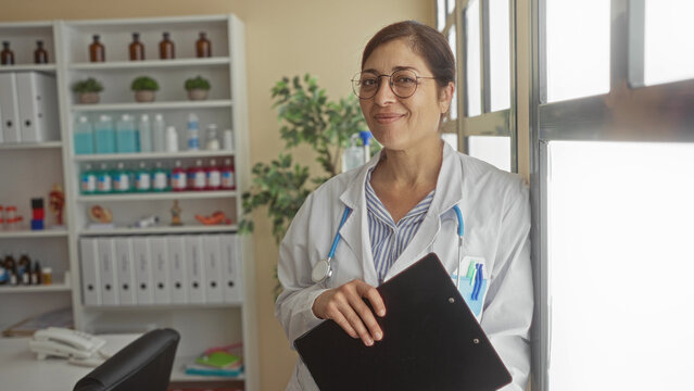 Middle aged hispanic woman doctor with stethoscope holds clipboard by window in clinic building; compassion dedication.