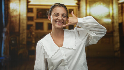 Obraz premium Woman smiling while making a call gesture with hand to ear inside ornate church building with chandeliers and paintings; playful invitation.