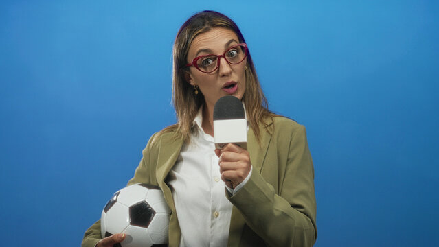 Woman holding microphone and soccer ball in blue studio speaking to camera; sports reporting confident.