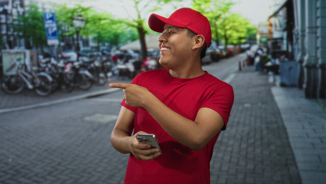 Man wearing red cap and shirt holding smartphone and pointing finger on busy city street with bicycles and buildings visible; happy delivery service. - Powered by Adobe