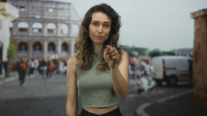 Woman pointing finger to temple in front of ancient coliseum building with blurred tourist crowd;...