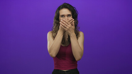 Young hispanic woman claps hands with open mouth wearing burgundy tank top and long curly hair in purple studio; surprise.