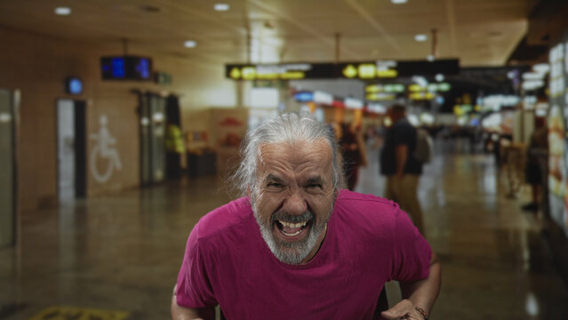 Man with clenched fists and hands cupped to mouth shouting in busy airport terminal, visible gray hair and beard, wearing magenta t shirt; joy celebration energy.