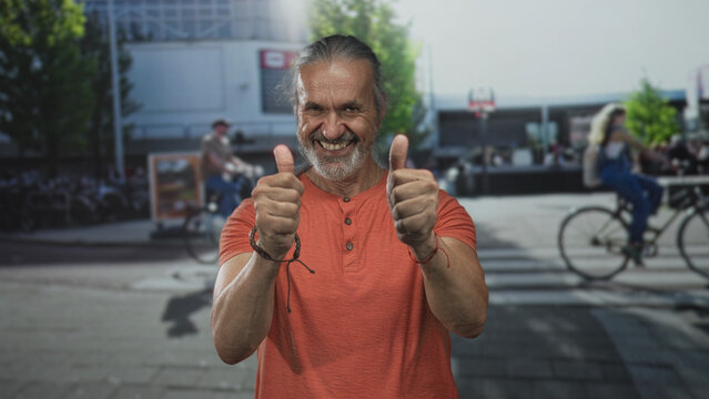 Man smiling and giving two thumbs up on a city street crosswalk in orange shirt and ponytail; happiness optimism.