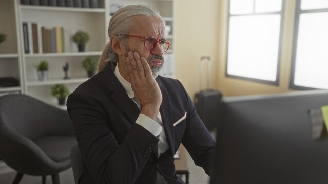 Man in suit with hand on cheek and toothache at office building desk computer, wearing red glasses and grey hair; work stress.